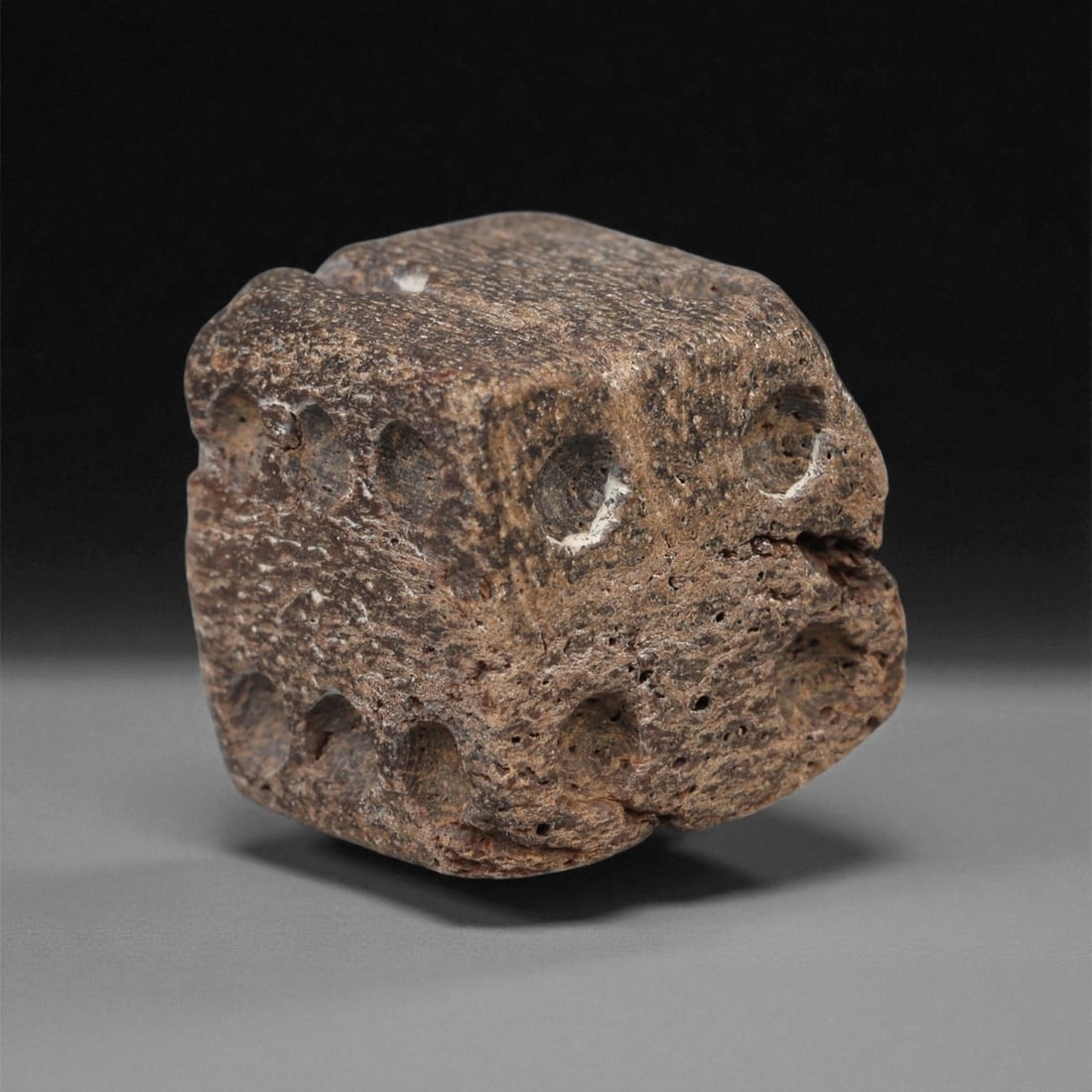 A Roman Bone Gaming Die, 1st-3rd Century CE, 68ZA: Carved from dense bone, the object is shaped as a small cube with slightly rounded edges. Each face bears a series of drilled circular pips arranged in standard numerical configurations. The surfaces 
