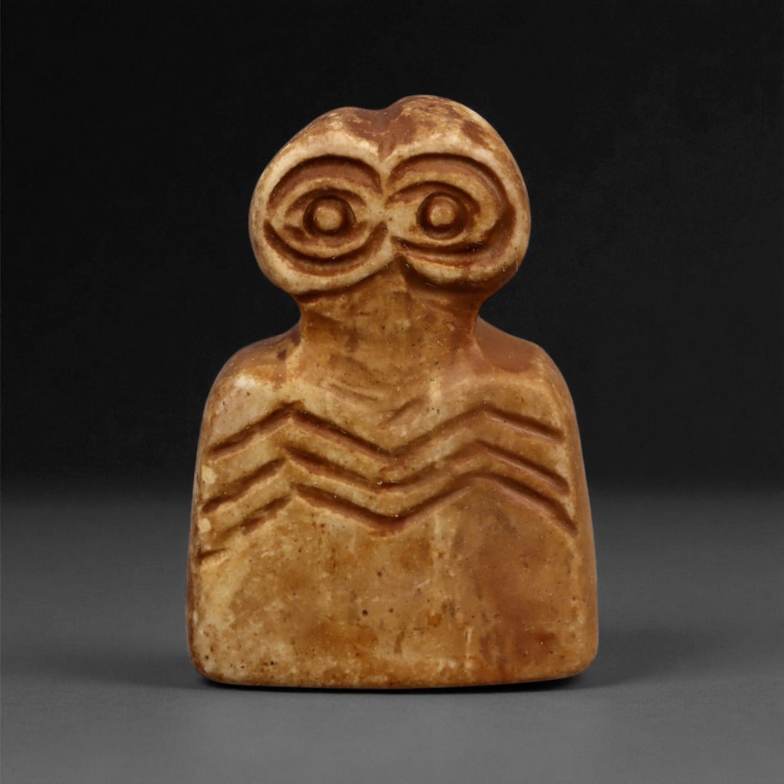 A Near Eastern Stone Eye Idol, 4th-3rd Millennium BCE, 54LA: Carved from light brown stone with darker surface toning, the figure has a broad lower body surmounted by a narrowed neck and an enlarged head. The eyes are rendered as two concentric recessed circles