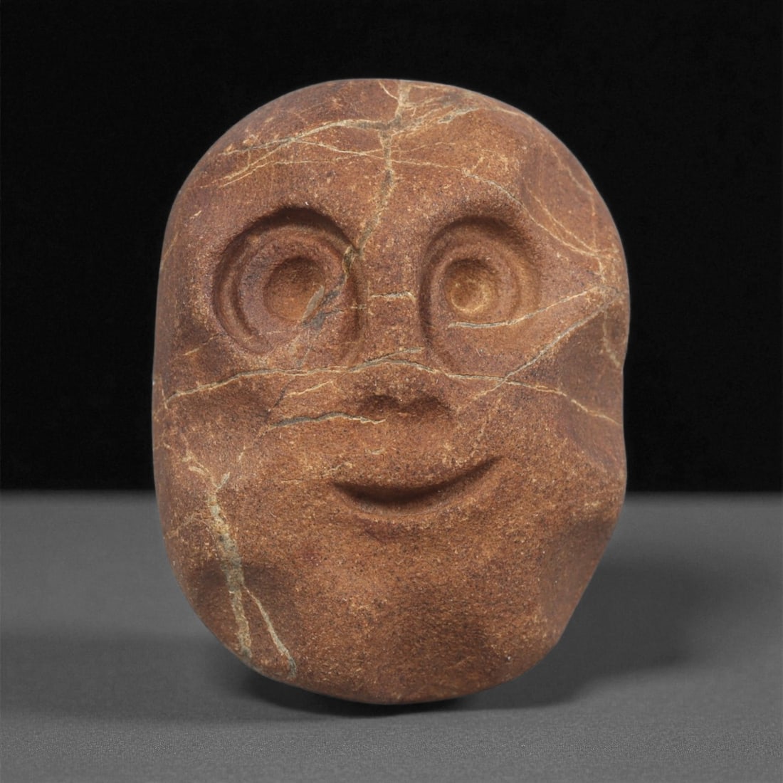 A Mediterranean Stone Maskette, 12th-17th Century, 100ZA: Carved from a reddish-brown stone with lighter veining across the surface, the maskette has a rounded, slightly tapering form. The facial features are rendered in shallow relief, including large conce