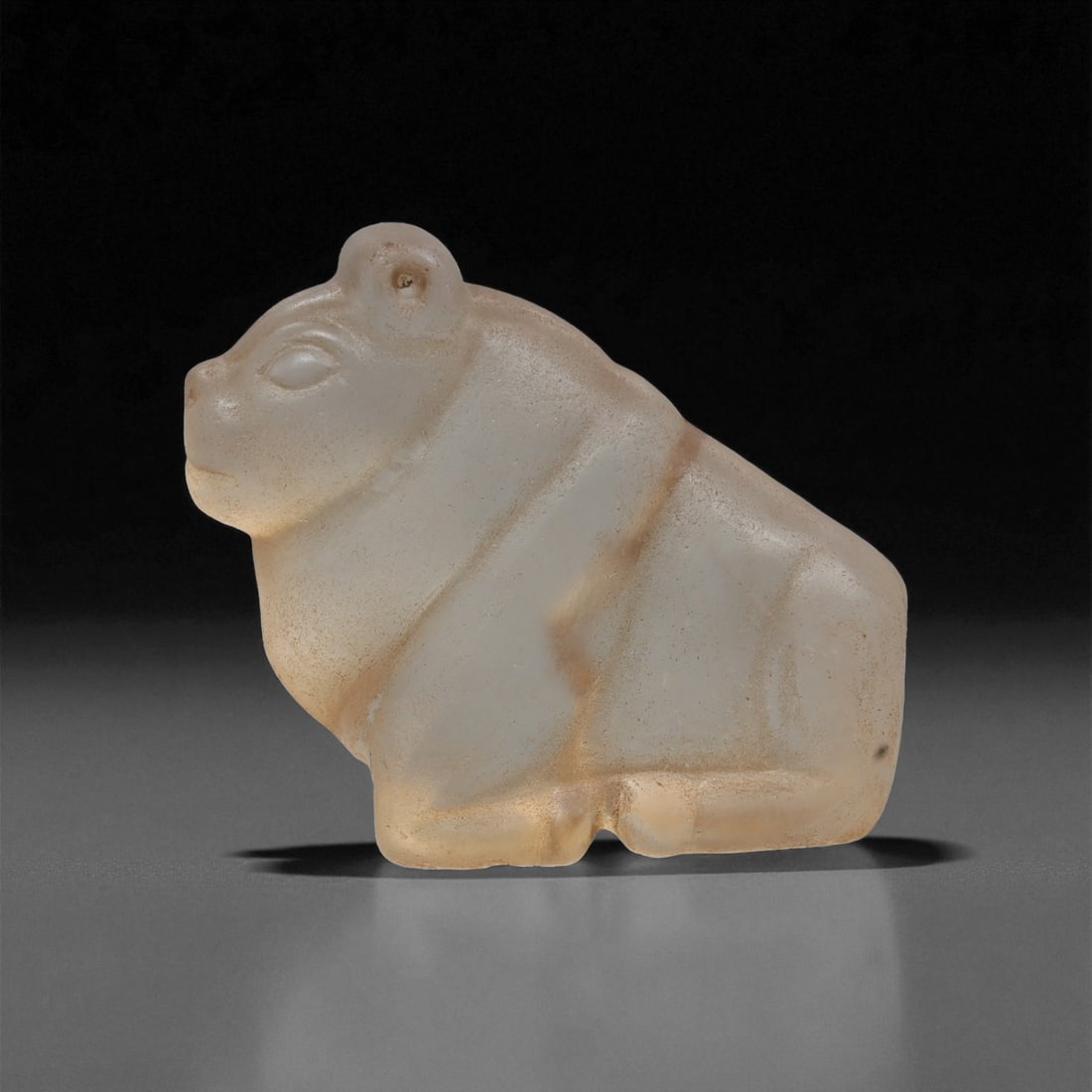 An Egyptian Coptic Glass Zoomorphic Figure, 4th-7th Century CE, 34ZB: Cast from translucent pale glass with minor surface weathering, the figure is shaped as a compact seated animal with a rounded body, short limbs, and a slightly raised head. The eyes and ears are deli
