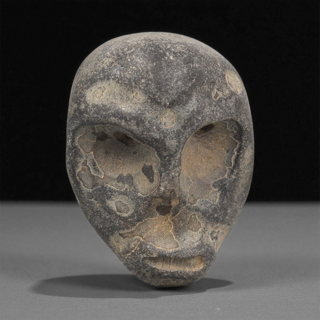 A Mediterranean Stone Maskette, 12th-17th Century, 6LC: Carved from a dark grey stone with lighter surface inclusions and patches of differential weathering, the maskette is shaped with an ovoid outline tapering to a narrow chin. The facial features are re
