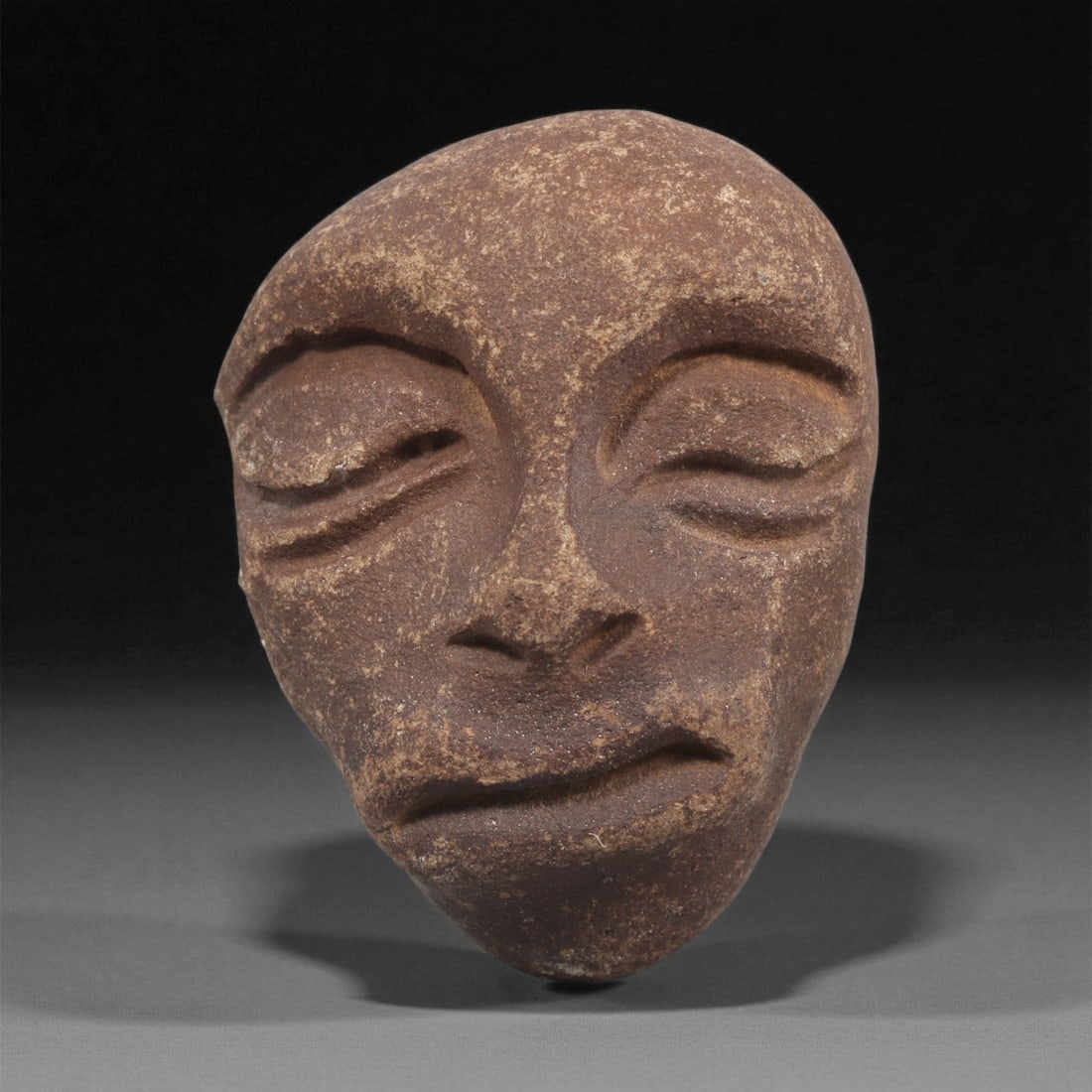A Mediterranean Stone Maskette, 12th-17th Century, 82LC: Carved from a dark brown stone with fine granular surface texture, the maskette is shaped with a rounded upper register tapering to a narrow chin. The facial features are rendered in shallow relief, i