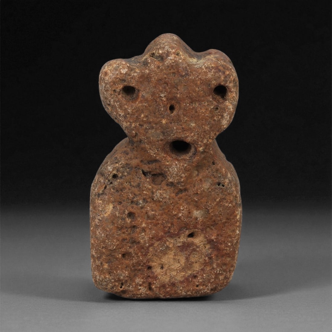 A Near Eastern Stone Anthropomorphic Figurine, 3rd-2nd Millennium BCE, 80LB: Carved from coarse, reddish-brown stone with scattered surface pitting, the figurine is shaped with a broad lower section and a distinctive tri-lobed head. The facial area is marked by three drilled a