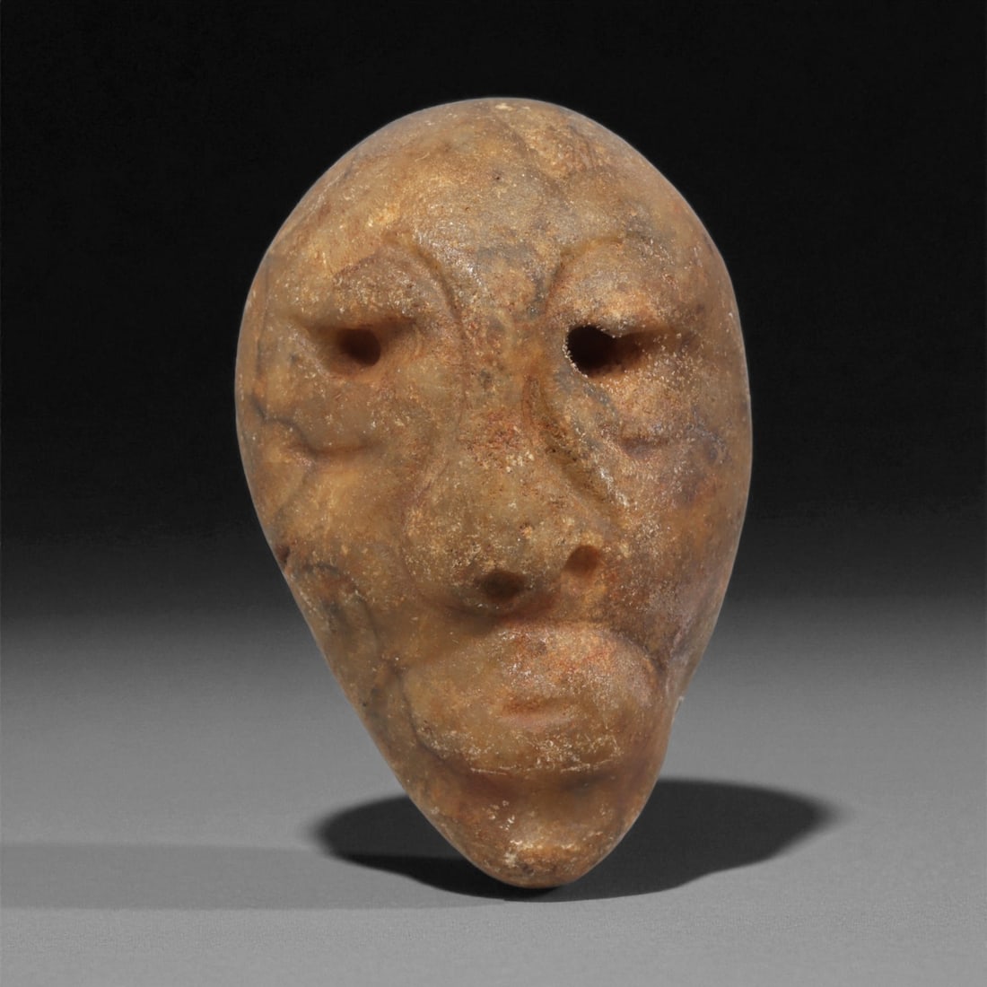 A Mediterranean Stone Maskette, 12th-17th Century, 88LC: Carved from a light brown stone with scattered surface inclusions, the maskette is shaped with an elongated oval outline tapering toward the chin. The facial features are rendered in shallow relief, i