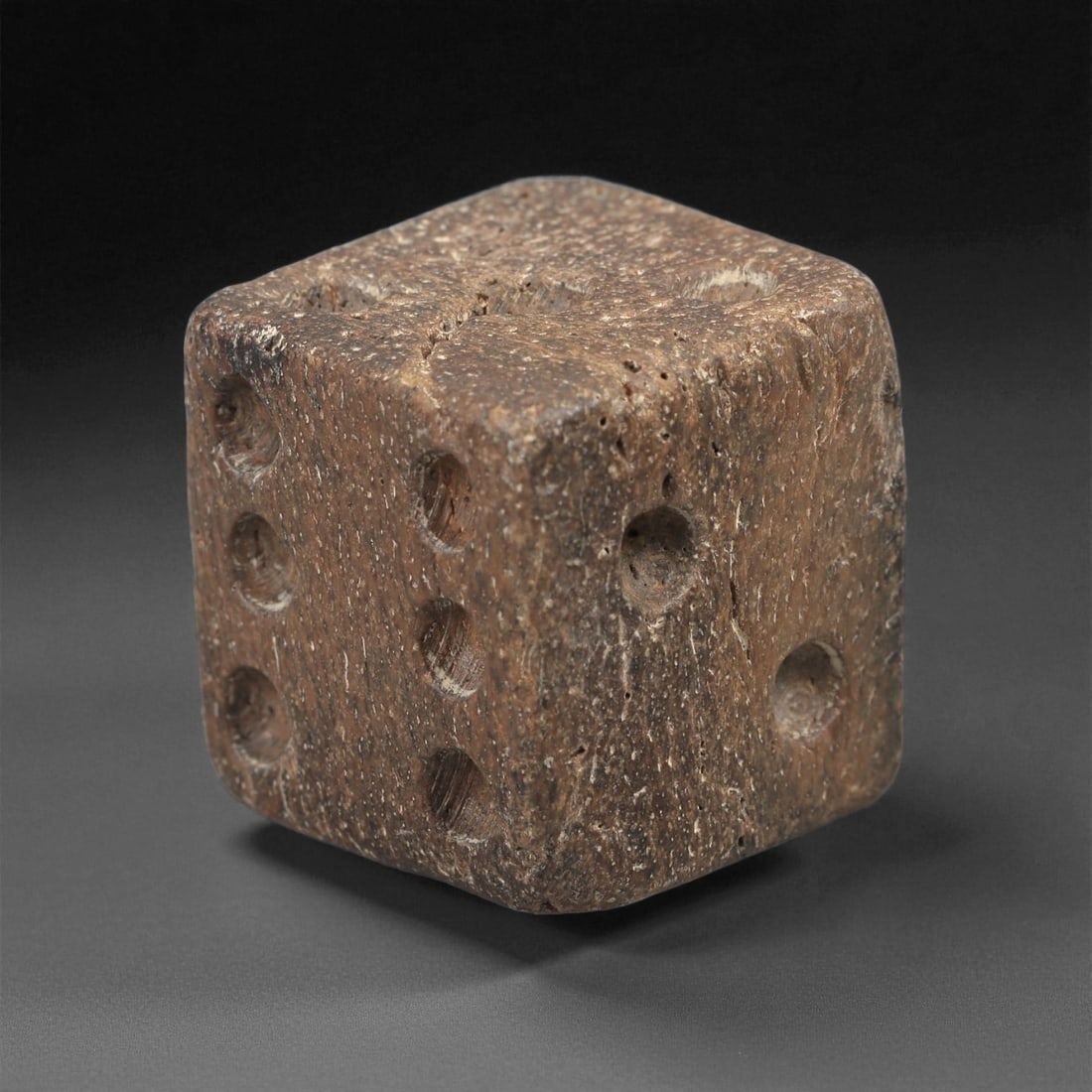 A Roman Bone Gaming Die, 1st-3rd Century CE, 61LD: Carved from light brown bone with a porous, naturally textured surface, the cubic die bears circular recessed pips on each face arranged in standard numerical configurations. The edges are slightly ro