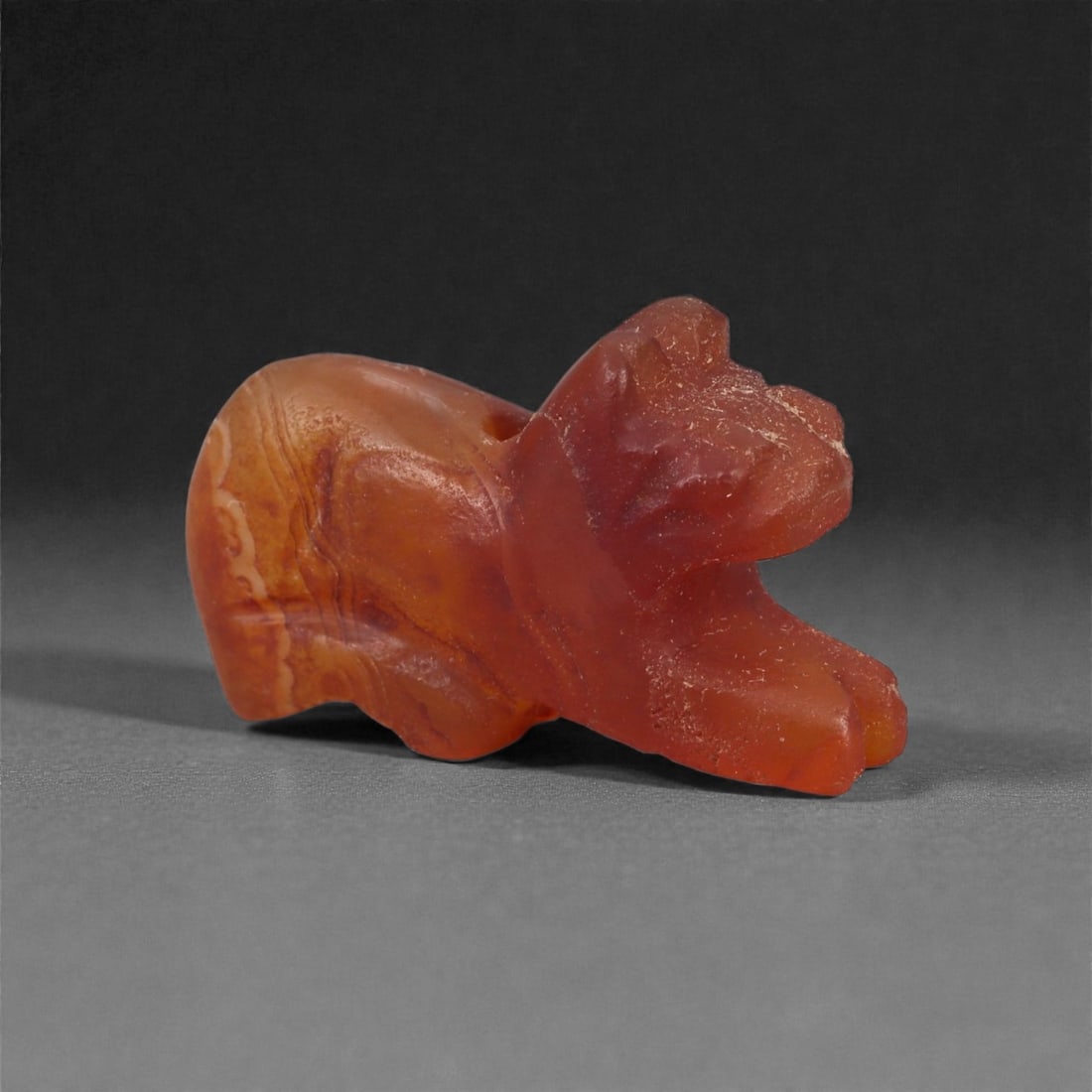 A Near Eastern Carnelian Lion Amulet, 1st Millennium BCE, 75LG: Carved from translucent orange carnelian with natural internal banding, the figure is modeled in the round as a recumbent animal with a compact body and a slightly raised head. The facial features are