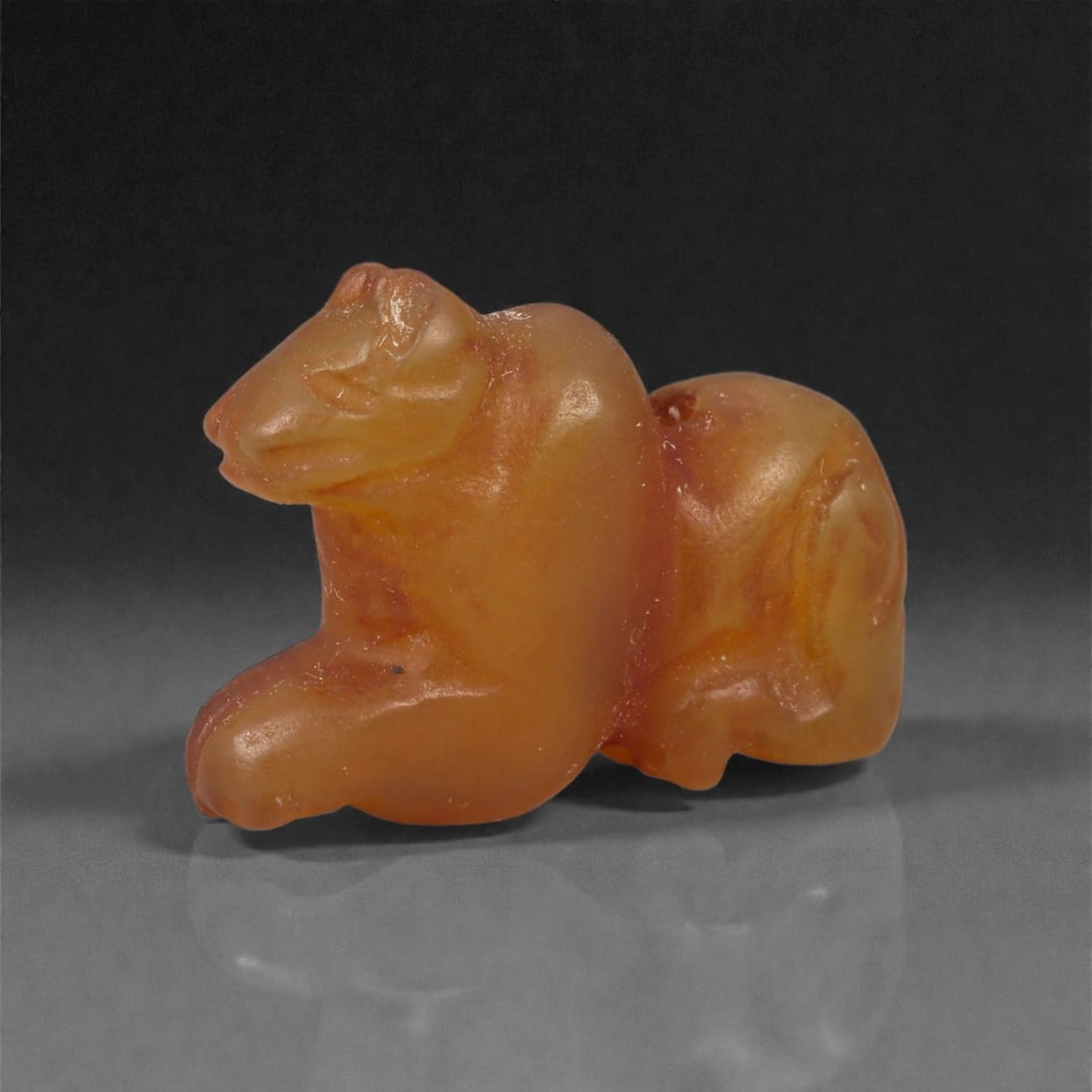 A Near Eastern Carnelian Lion Amulet, 1st Millennium BCE, 76LG: Carved from translucent orange carnelian, the amulet depicts a recumbent lion rendered in a compact, rounded form. The head is defined by incised lines indicating the eyes and muzzle, while the forele