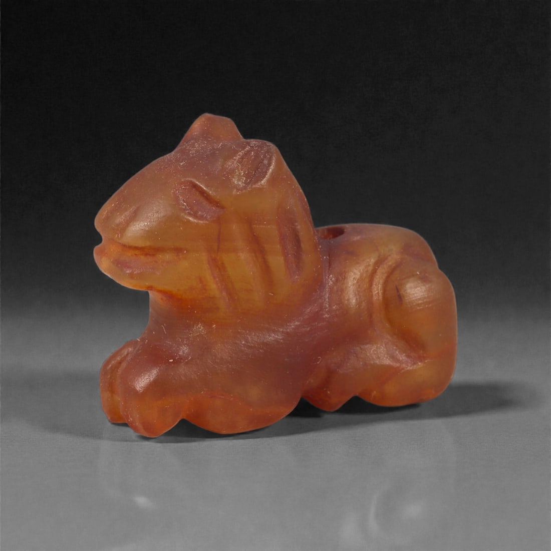 A Near Eastern Carnelian Lion Amulet, 1st Millennium BCE, 89LG: Carved from translucent orange carnelian with natural internal banding, the figure is modeled in the round as a recumbent animal with a compact body and a slightly raised head. The facial features are