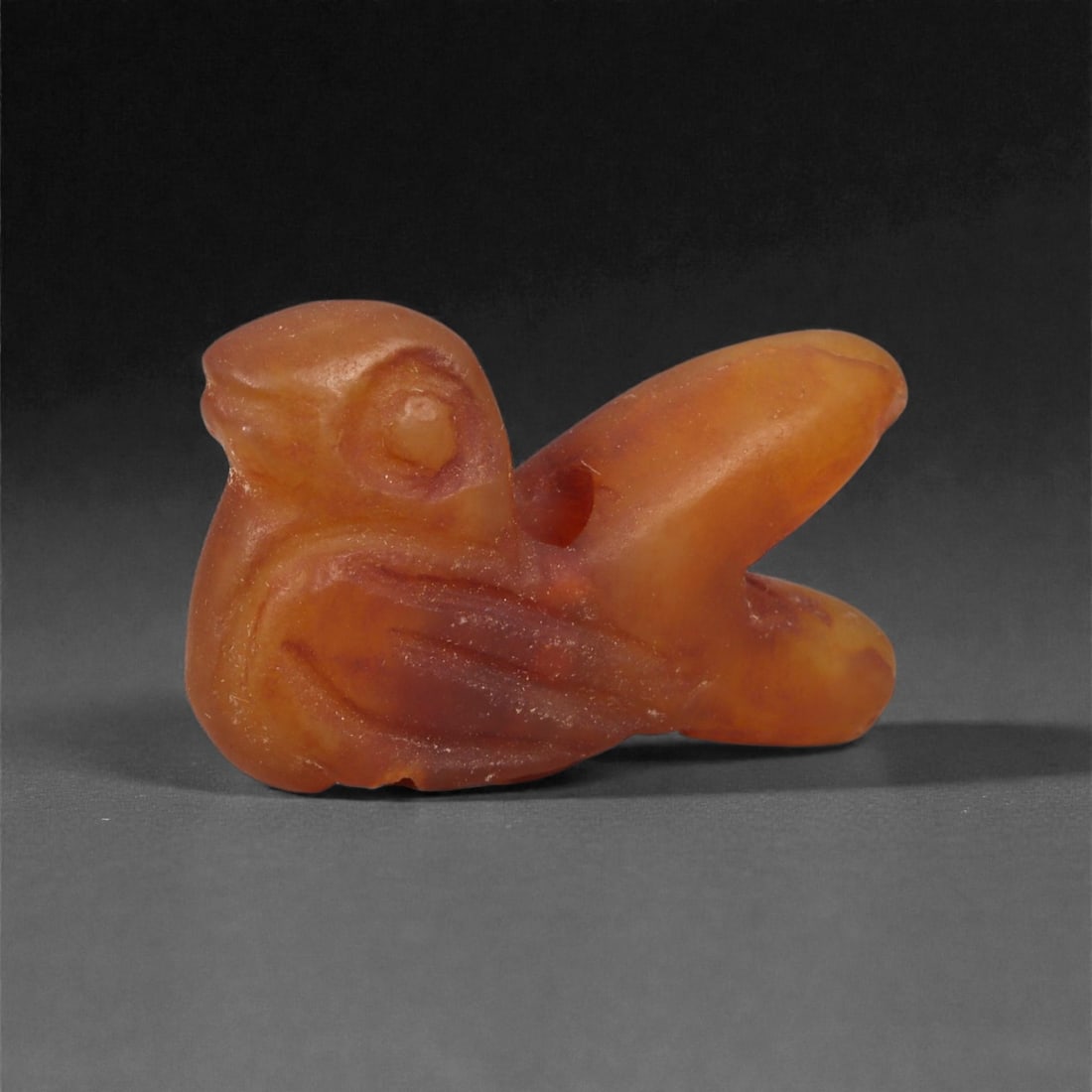A Near Eastern Carnelian Bird Amulet, 1st Millennium BCE, 74LG: Carved from orange-red carnelian with natural internal color zoning, the amulet depicts a stylized bird with a compact body and rounded head. The wings are indicated through shallow linear carving alo