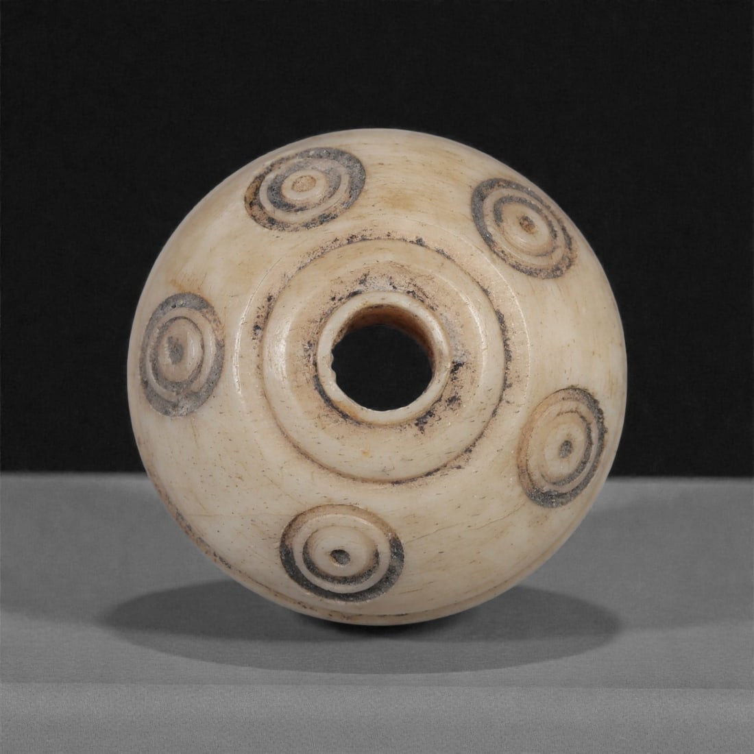 A Near Eastern Carved Bone Spindle Whorl, 1st Millennium BCE, 96LH: Carved from a light cream-colored bone with fine surface patination, the whorl is shaped with a domed profile and a central perforation surrounded by a raised circular ridge. Five concentric ring-and-
