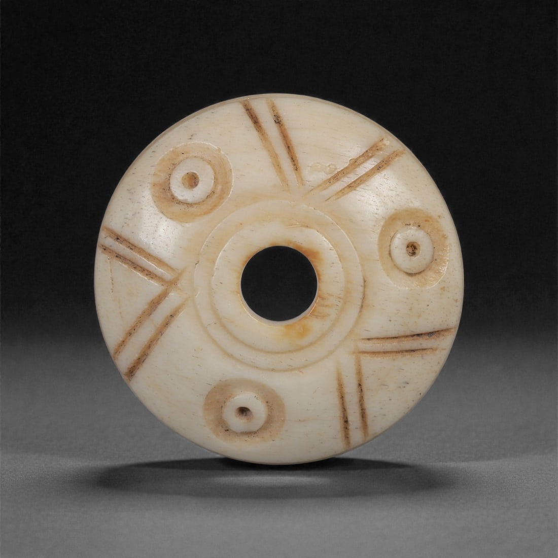A Near Eastern Carved Bone Spindle Whorl, 1st Millennium BCE, 98LH: Carved from a fine, pale bone with smooth creamy patination, the whorl is shaped with a flat-domed profile and a neatly drilled central perforation bordered by a raised circular ridge. The upper surfa