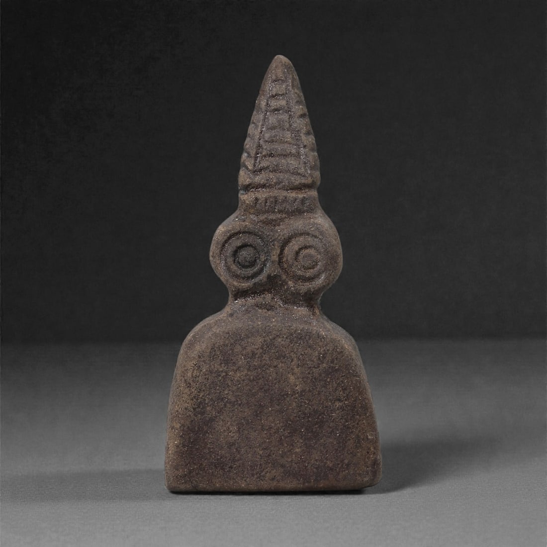 A Near Eastern Stone Eye Idol with Conical Headdress, 4th-3rd Millennium BCE, 61LH: Carved from a dark brown stone with a fine, evenly worn surface, the figure is shaped with a broad, squared lower body and a narrow neck supporting a twin-lobed head. The eyes are rendered as recessed