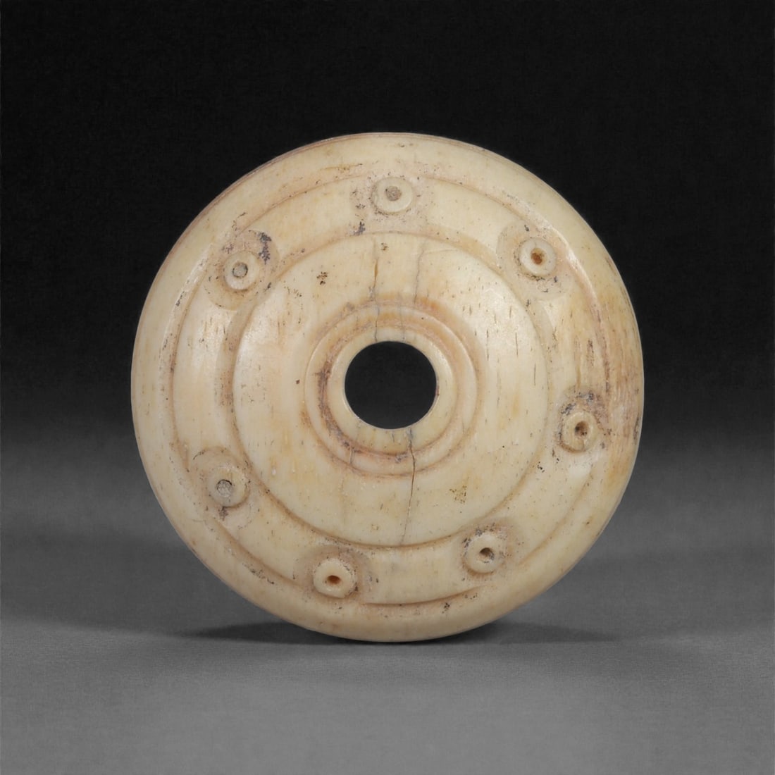 A Near Eastern Carved Bone Spindle Whorl, 1st Millennium BCE, 100LH: Carved from a light cream-colored bone with smooth surface patination, the spindle whorl is formed with a domed profile and a central perforation framed by a raised circular ridge. The upper surface b