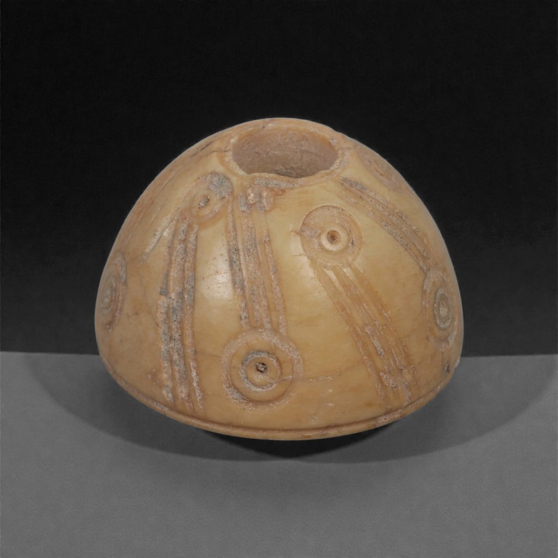 A Near Eastern Carved Bone Spindle Whorl, 1st Millennium BCE, 93LH: Carved from a light brown bone with smooth surface patination, the object is shaped as a domed spindle whorl with a broad, slightly flattened base and a central perforation. The exterior bears a serie