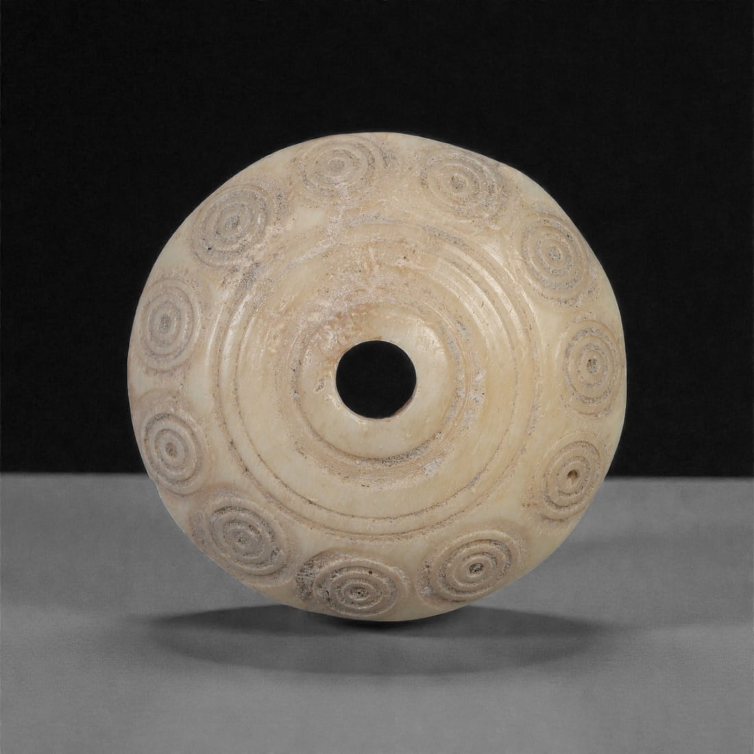 A Near Eastern Carved Bone Spindle Whorl, 1st Millennium BCE-1st Millennium CE, 1LJ: Carved from a light cream-colored bone with fine surface patination, the whorl is shaped with a domed profile and a central perforation bordered by multiple concentric incised rings. Around the outer 
