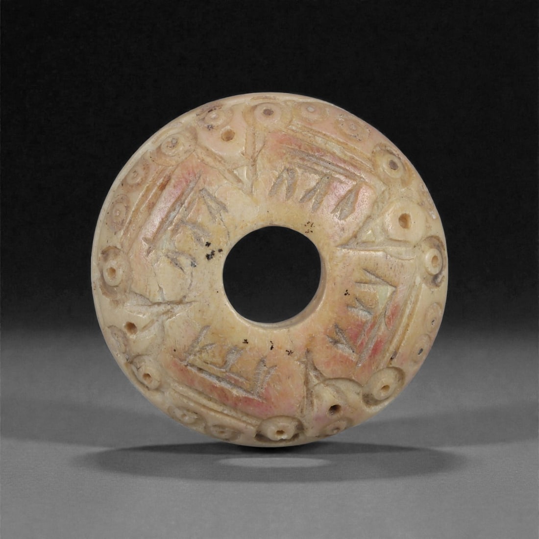 A Near Eastern Carved Bone Spindle Whorl, 1st Millennium BCE, 97LH: Carved from a light cream-colored bone with areas of reddish surface staining, the whorl is shaped with a gently domed profile and a central perforation. The upper surface carries an irregular arrange