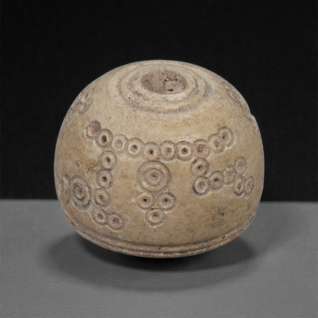 A Near Eastern Carved Bone Spindle Whorl, 1st Millennium BCE, 94LH: Carved from a warm beige-toned bone with areas of mineral accretion, the whorl is shaped with a rounded, slightly domed form and a central perforation framed by concentric incised rings. The surface i