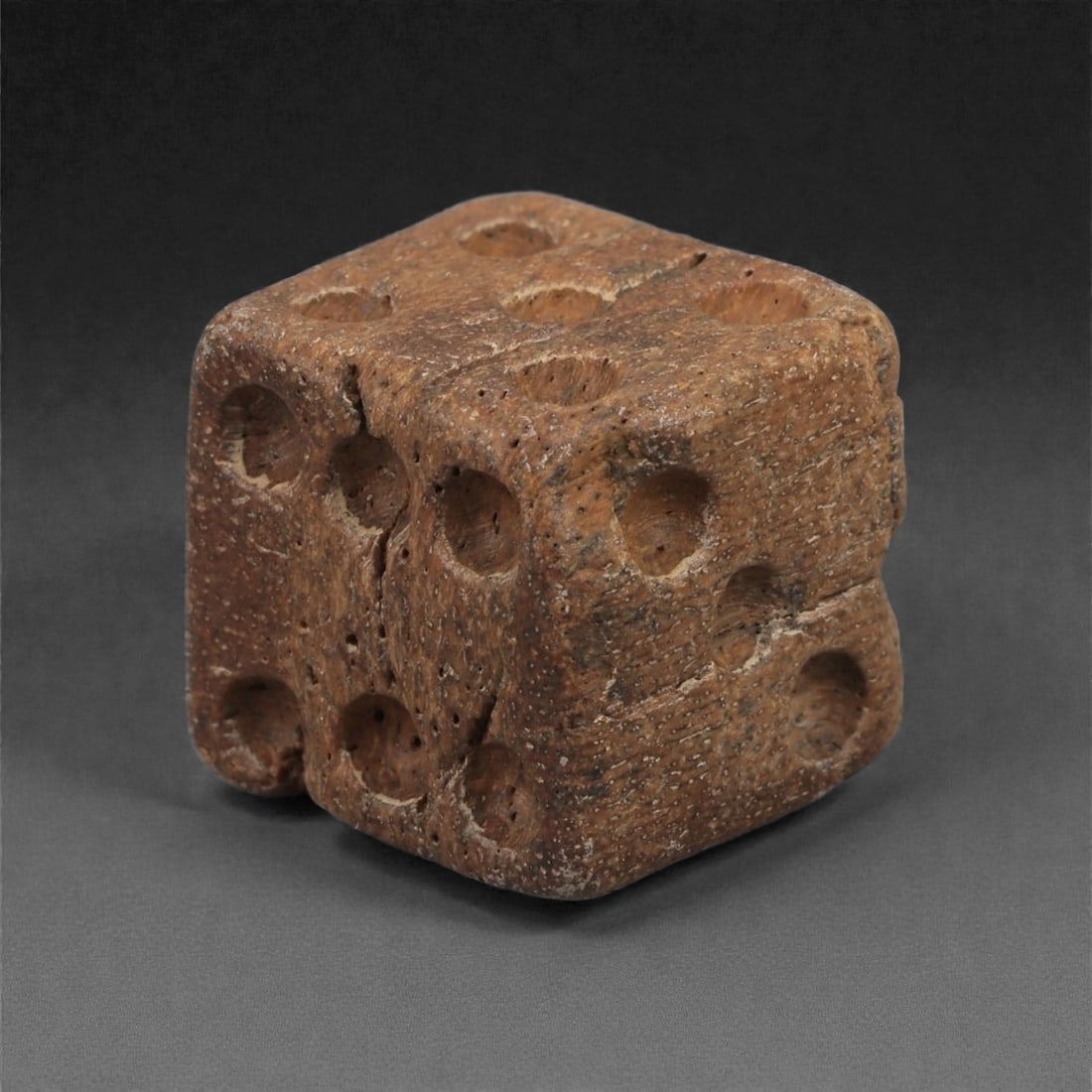 A Roman Bone Gaming Die, 1st-3rd Century CE, 1.5cm, 57LG: Carved from dense bone, the object is shaped as a small cube with slightly rounded edges. Each face bears a series of drilled circular pips arranged in standard numerical configurations. The surfaces 