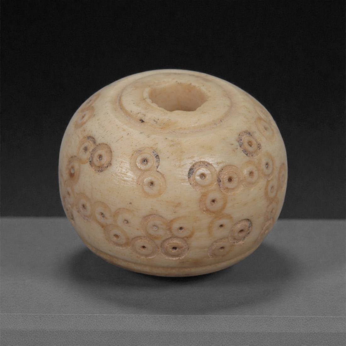 A Near Eastern Carved Bone Spindle Whorl, 1st Millennium BCE, 95LH: Carved from a light cream-colored bone with smooth surface patination, the whorl is shaped with a rounded, slightly flattened globular profile and a central perforation bordered by a shallow recessed 