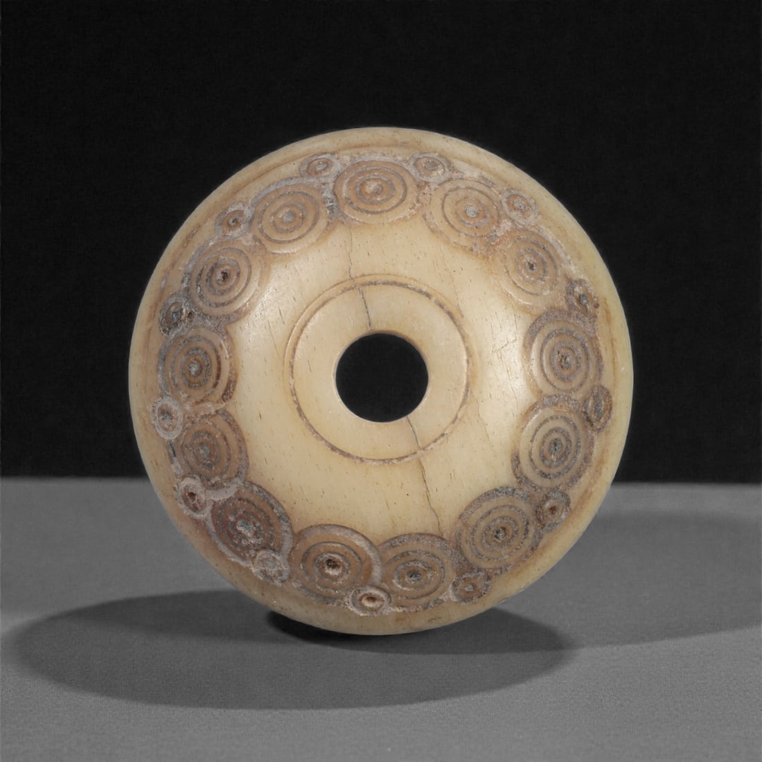 A Near Eastern Carved Bone Spindle Whorl, 1st Millennium BCE, 99LH: Carved from a light cream-colored bone with fine surface patination, the object is shaped as a domed spindle whorl with a central perforation. The upper surface is encircled by a continuous band of co
