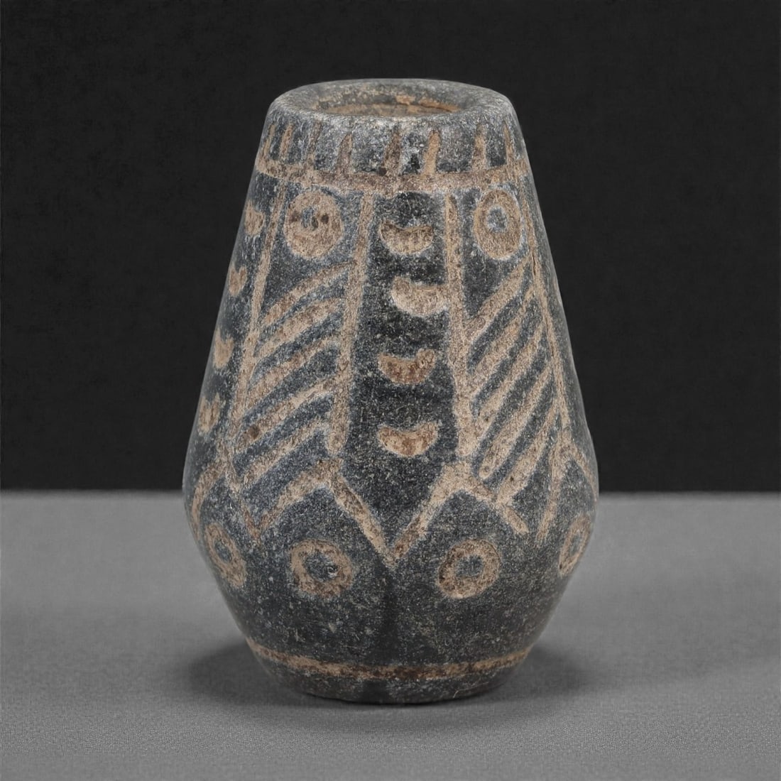 A Near Eastern Basalt Bead, 2nd-1st Millennium BCE, 76LJ: Carved from dark basalt, the bead is shaped with a tapering conical body and a flat top and base. The upper register bears vertically incised strokes along the rim and ring-and-dot motifs framed withi