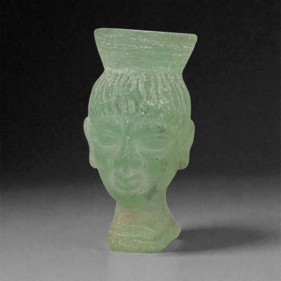 A Roman Glass Bust, 1st-3rd Century CE, 7LN: Cast in pale green glass with a matte, weathered surface, the bust depicts a figure with a rounded face, almond-shaped eyes, a straight nose, and a small mouth. The hair is rendered with vertical stri