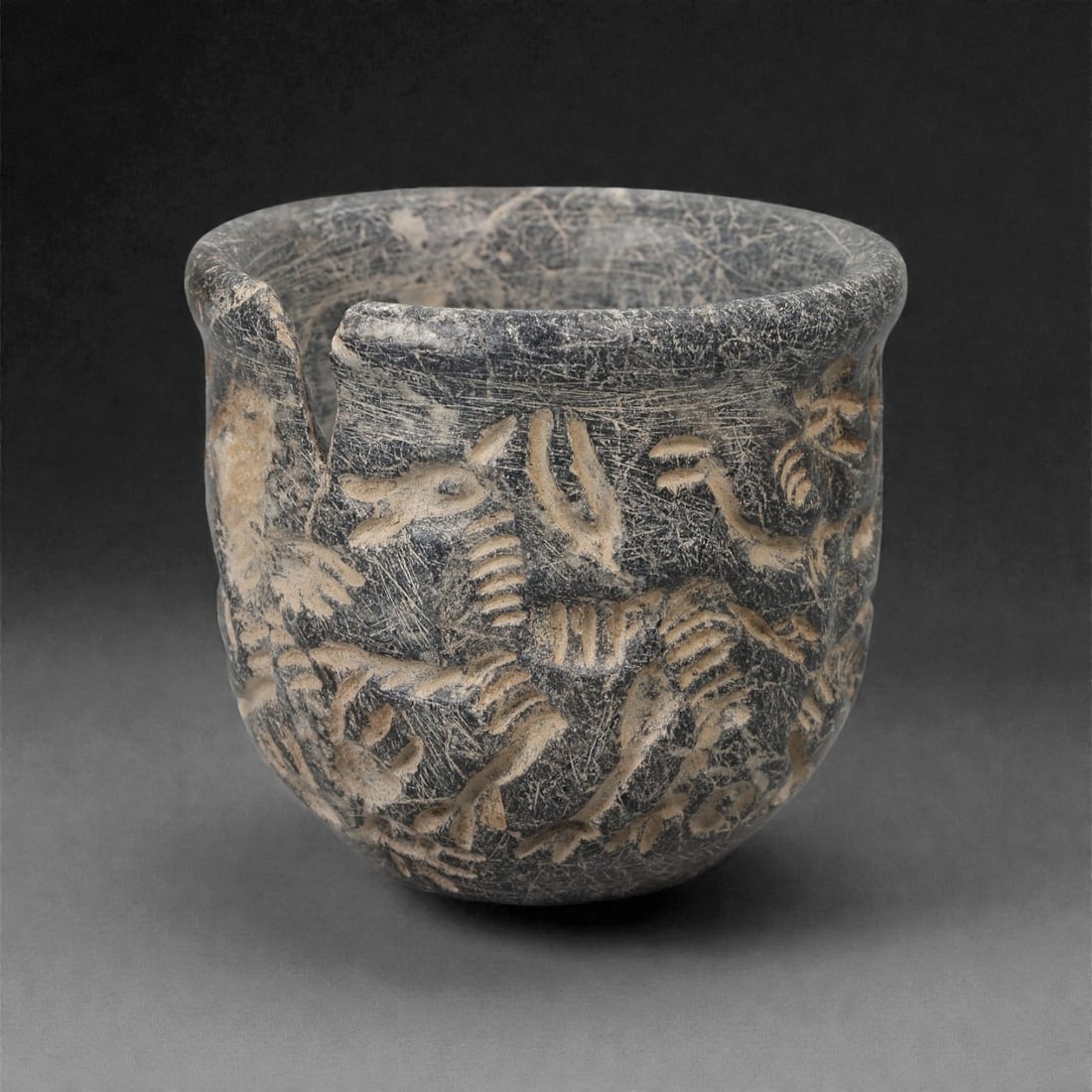 A Jiroft Steatite Cup with Animal Reliefs, 3rd Millennium BCE, 52LK: Carved from dark grey steatite with lighter inclusions, the cup has straight walls rising to a slightly thickened rim. The exterior is decorated in low relief with a continuous frieze of stylized anim