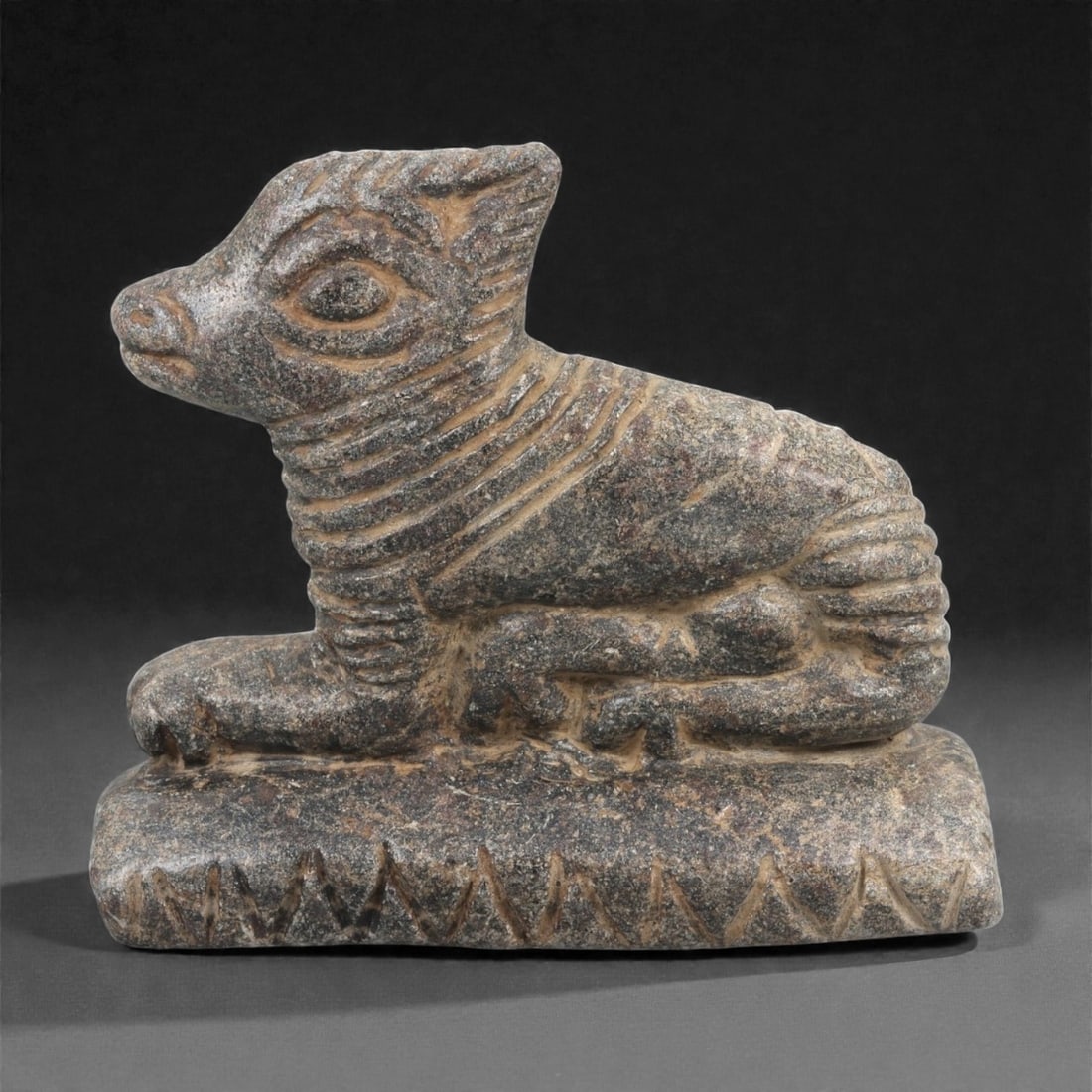 A Mesopotamian Schist Recumbent Calf, Early 2nd Millennium BCE, 64LJ: Carved from dark grey schist, the figure shows a calf in a recumbent position with the forelegs extended and the hind legs folded beneath the body. The head is shaped with large recessed oval eyes, a 