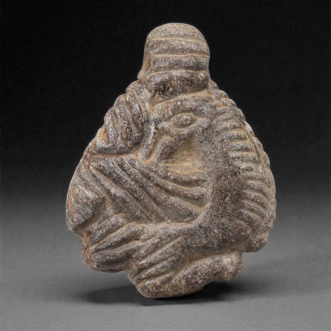 A Syro-Hittite Schist Bird Amulet, Early Iron Age, 11th-9th Century BCE, 89LJ: Carved from grey-brown schist, the amulet takes the form of a compact plaque surmounted by a rounded, horizontally banded knob serving as an integrated suspension element. The front displays a bird in