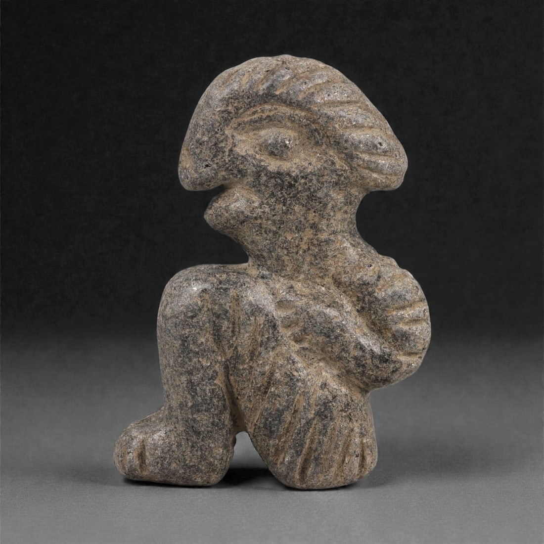 A Syro-Hittite Schist Anthropomorphic Figurine, Early Iron Age, 11th-9th Century BCE, 69LJ: Carved from grey schist, the figure is shown seated with one knee raised and the opposite leg folded beneath the body. The head is rendered with an elongated cranial profile, a prominent recessed eye,