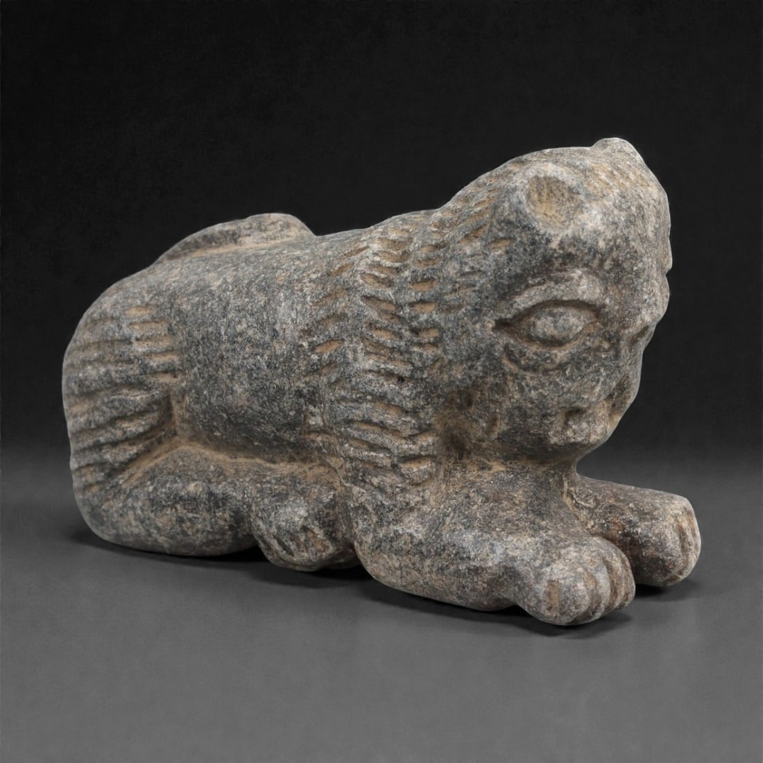 A Syro-Anatolian Schist Feline Figure, Early to Mid-2nd Millennium BCE, 85LJ: Carved from mottled grey schist, the figure represents a recumbent feline with the forepaws extended forward and the hind limbs folded beneath the body. The head is rounded with recessed circular eyes