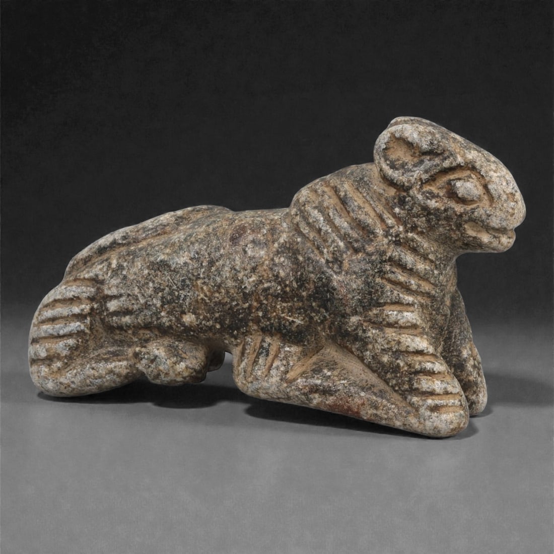 A Syro-Anatolian Schist Feline Figure, Early to Mid-2nd Millennium BCE, 57LJ: Carved from mottled grey schist, the figure depicts a recumbent feline with the forelegs extended forward and the hind limbs folded beneath the body. The head is rounded with recessed circular eyes, a