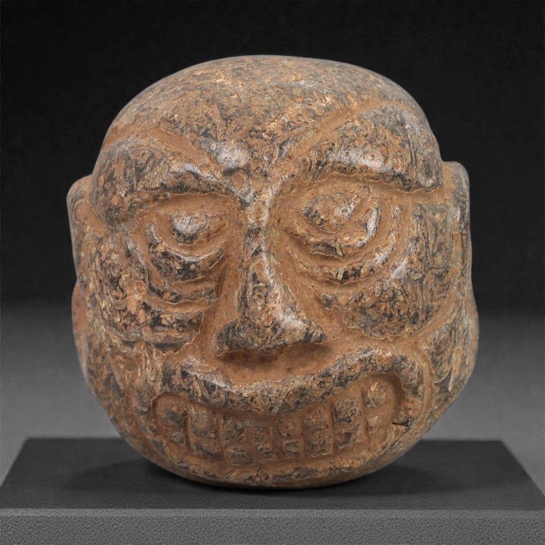 A Syro-Levantine Stone Anthropomorphic Maskette, Iron Age, c. 10th-8th Century BCE, 74LN: Carved from brown stone with darker surface mottling, the object forms a rounded anthropomorphic head with deeply recessed eyes framed by concentric incised arcs, a prominent straight nose, and a wide