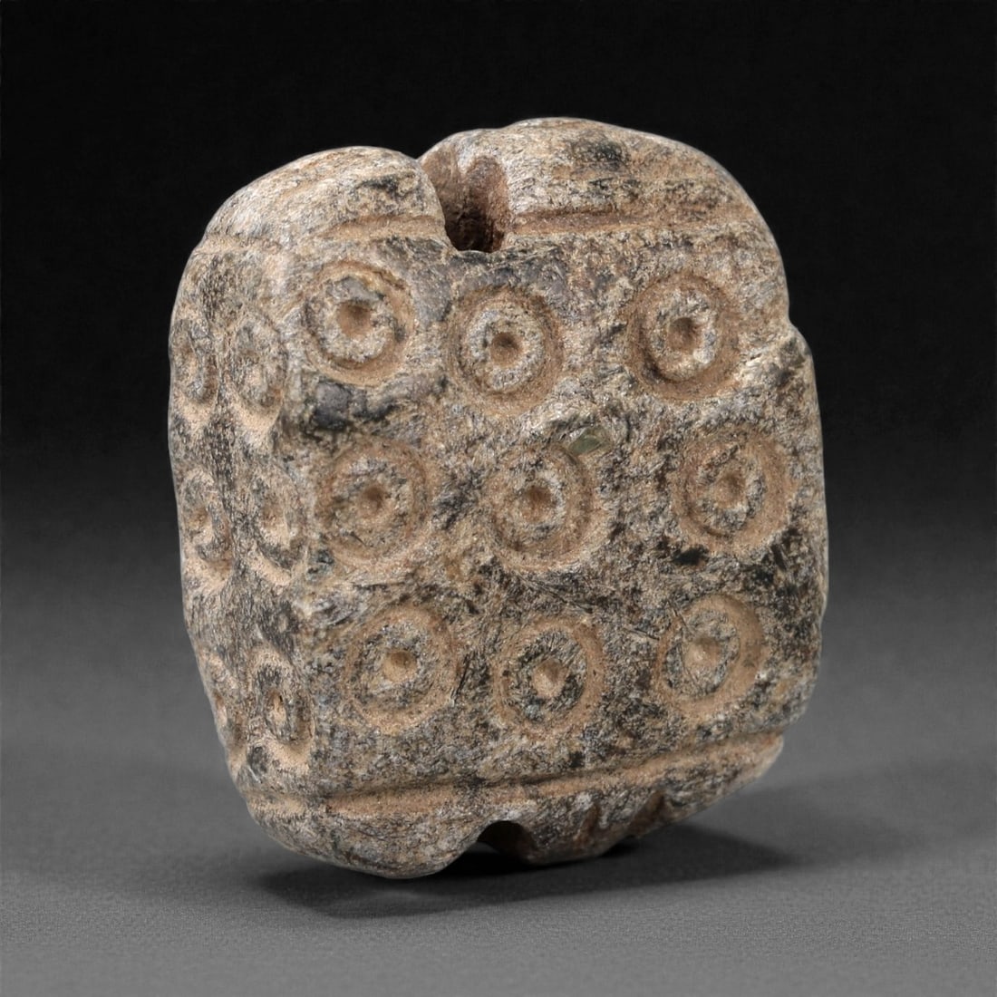 A Bactrian Stone Cylindrical Bead, Bronze Age, Circa 2200-1800 BCE, 61LN: Carved from mottled grey-brown stone, the bead is of short cylindrical form with a central perforation running vertically through the body. The surface is decorated with rows of drilled concentric cir