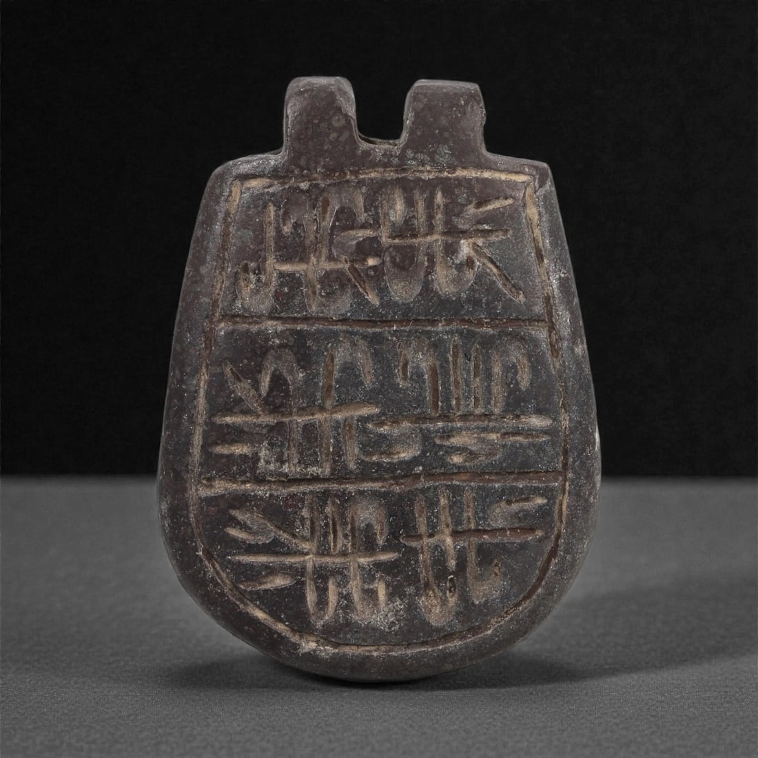 A Mesopotamian Steatite Plaque Amulet, Early Dynastic to Akkadian, c. 2600-2200 BCE, 91LN: Carved from dark grey steatite, the amulet is formed as a tapering plaque with a rounded lower edge and twin upright lugs at the top serving as an attachment feature. The front is divided into three h