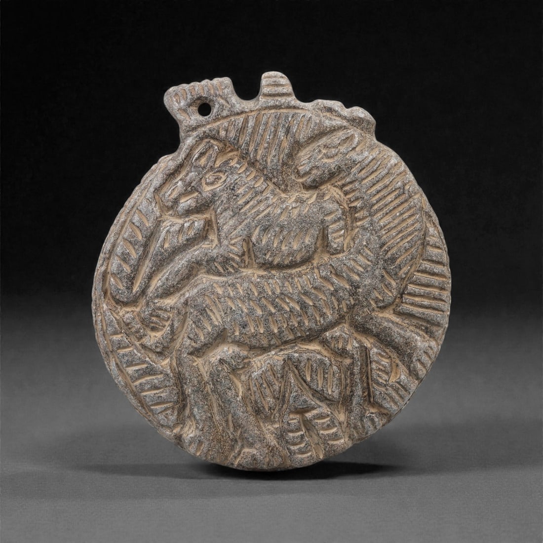 A Syro-Anatolian Schist Zoomorphic Amulet, Iron Age, Circa 9th-7th Century BCE, 92LN: Carved from grey schist, the circular plaque is worked in low relief with a pair of superimposed quadrupeds rendered in a compact composition. Both animals exhibit elongated bodies with incised hatchi