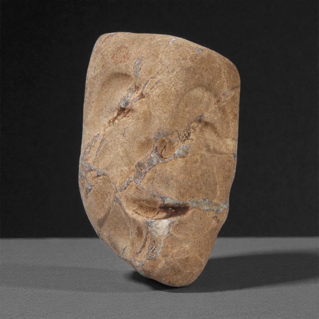 A Mediterranean Stone Maskette, 12th-17th Century, 48AD: Carved from a light brown stone with prominent natural fissures and surface veining, the maskette is formed with an asymmetrical outline that tapers toward the chin. The facial features are rendered i