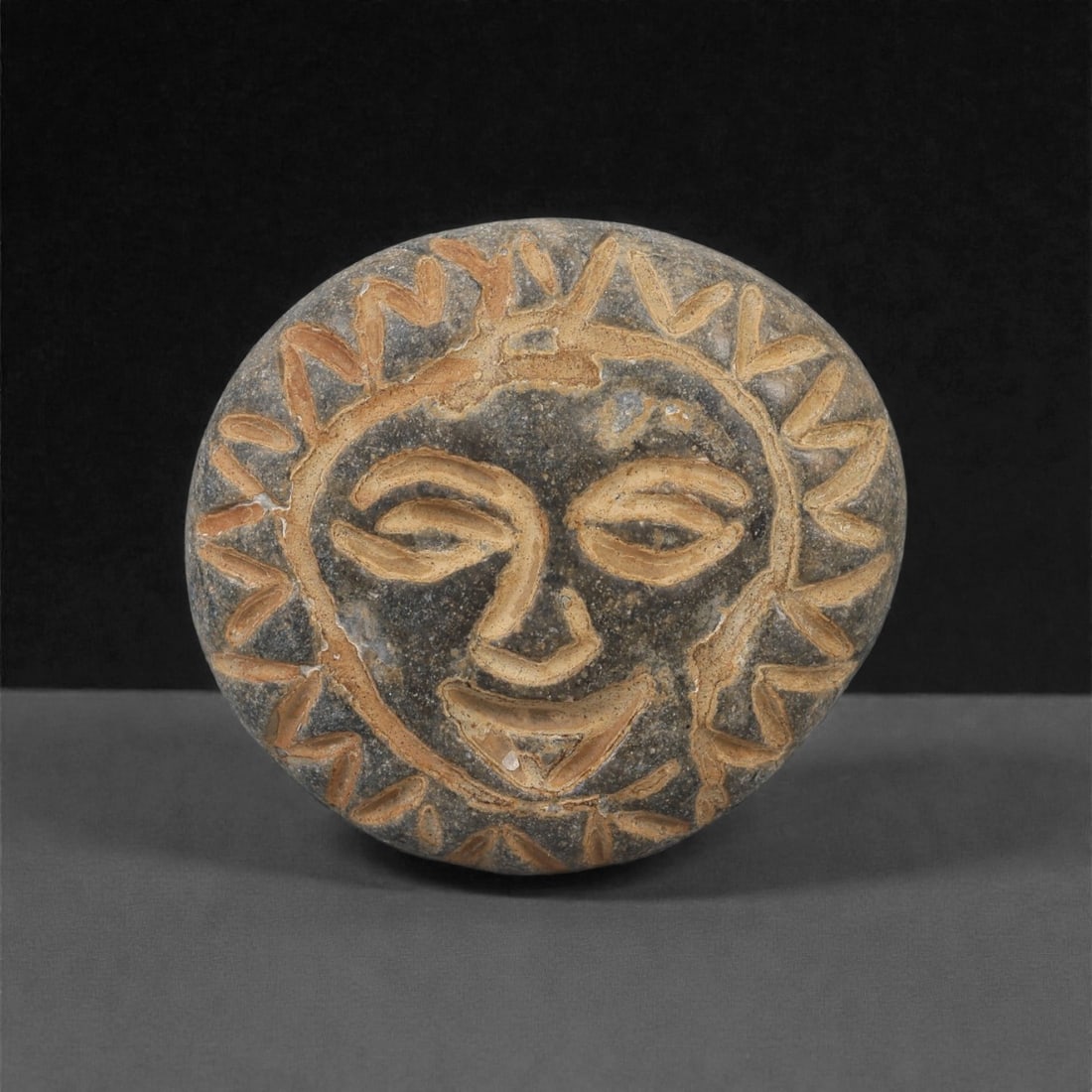 A Mediterranean Stone Maskette, 12th-17th Century, 57AD: Carved from a dark grey stone with lighter inclusions, the maskette is shaped with a rounded outline and flat reverse. The face is rendered in shallow incision, including almond-shaped eyes, a linear 