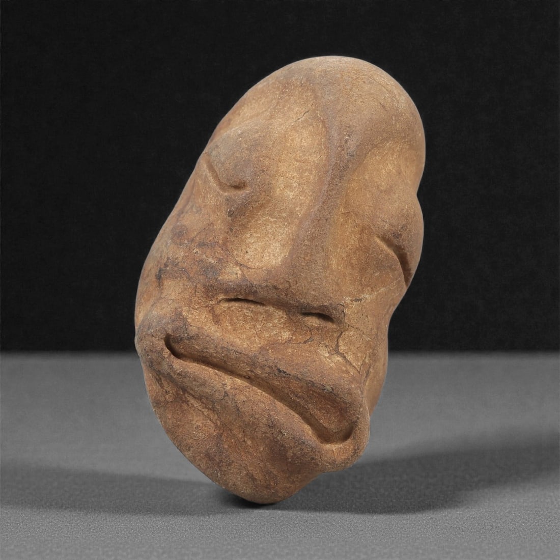 A Mediterranean Stone Maskette, 12th-17th Century, 29AD: Carved from a light brown stone with fine surface granularity, the maskette is shaped as an elongated face with a markedly asymmetrical contour. The eyes are indicated by recessed, closed lids carved 
