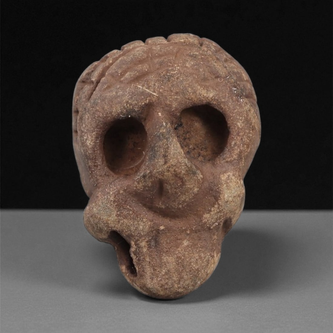 A Mediterranean Stone Maskette, 12th-17th Century, 12AD: Carved from a reddish-brown stone with granular surface texture, the maskette is shaped as a compact head with a rounded upper register marked by incised cross-hatched lines. The eyes are formed as tw