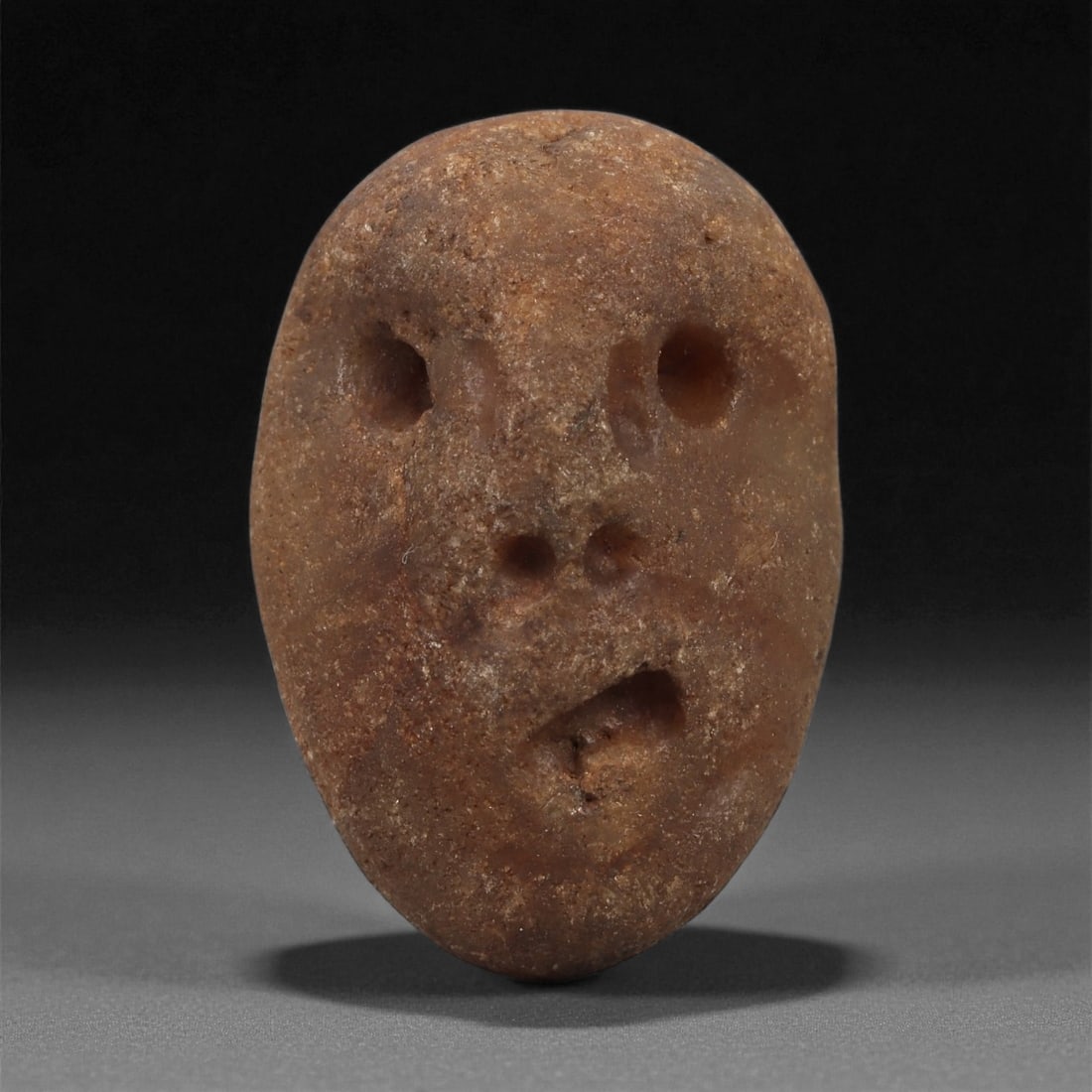 A Mediterranean Stone Maskette, 12th-17th Century, 88AB: Carved from a warm reddish-brown stone with a naturally weathered, granular surface, this small maskette presents an elongated oval face with minimal but expressive features. The eyes are formed as tw
