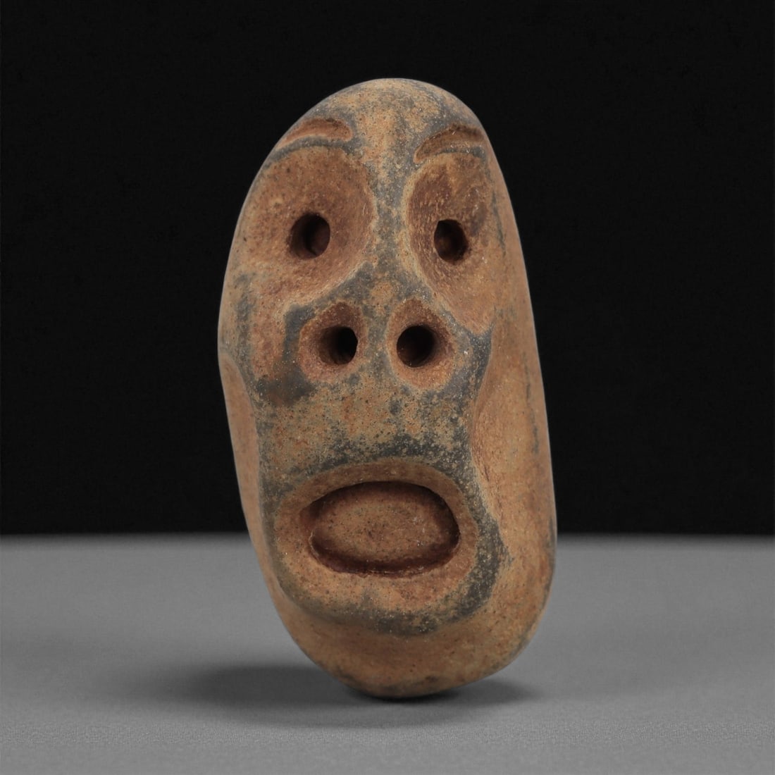 A Mediterranean Stone Maskette, 12th-17th Century, 2AD: Carved from a light brown stone with darker surface accretions, the maskette is formed as an elongated oval face with a slightly asymmetrical contour. The eyes are represented by two deep circular per