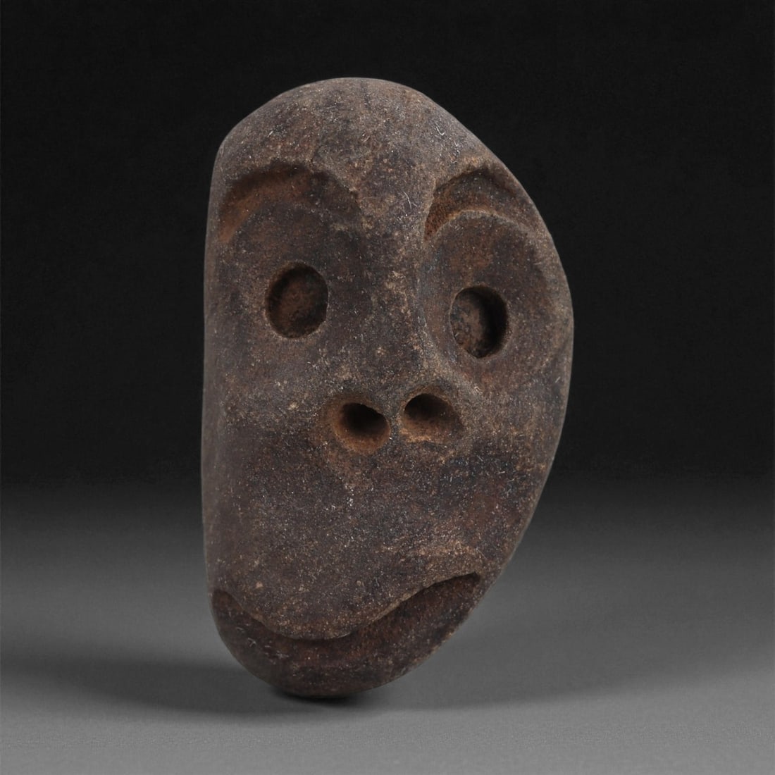 A Mediterranean Stone Maskette, 12th-17th Century, 8AD: Carved from a dark brown stone with a compact, slightly irregular surface, the maskette presents an elongated facial form with a subtly asymmetrical contour. The eyes are represented by deep, circular