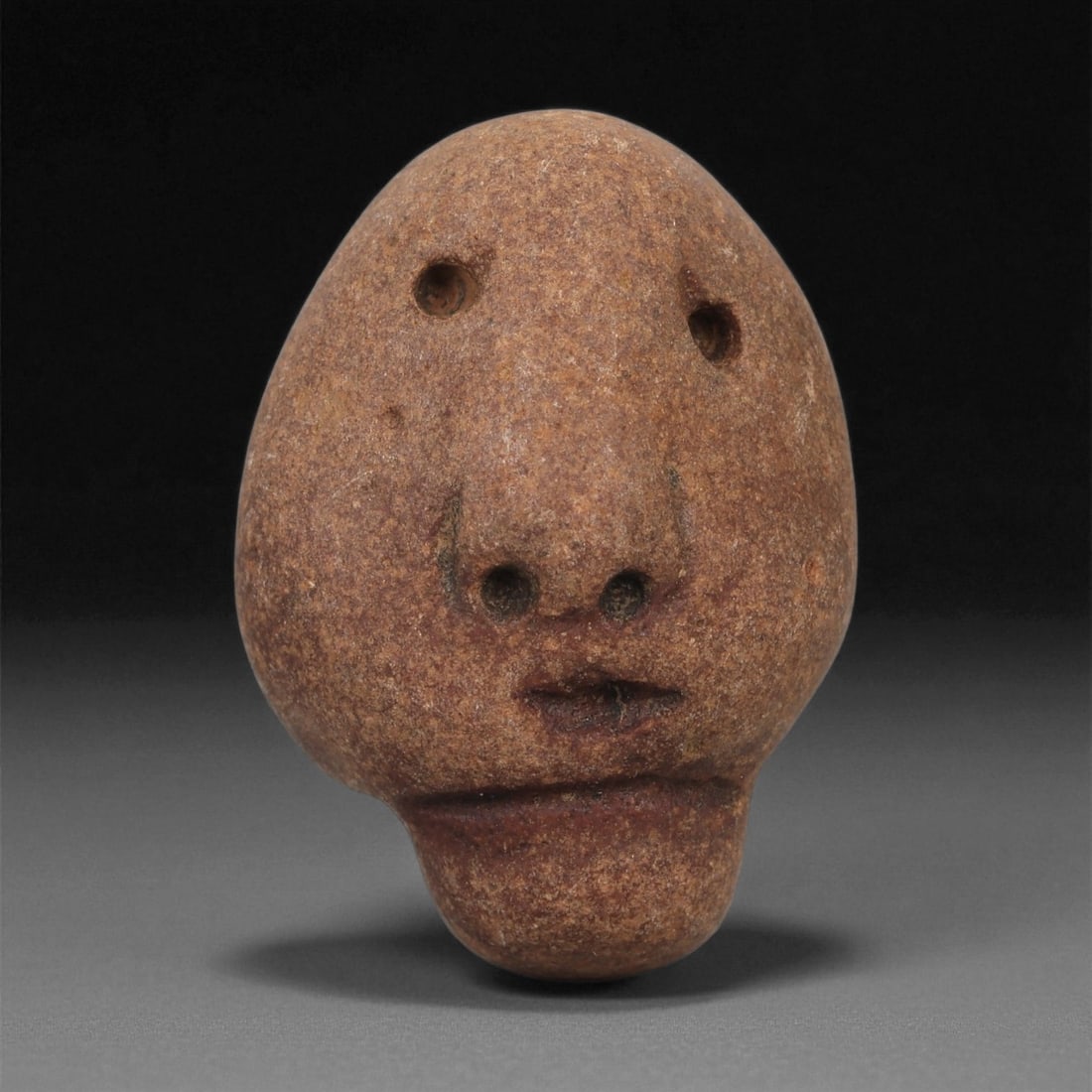 A Mediterranean Stone Maskette, 12th-17th Century, 98AB: Carved from a reddish-brown stone with fine granular texture, the maskette is shaped as an ovate head tapering toward an elongated chin. The eyes are represented by two drilled circular apertures plac