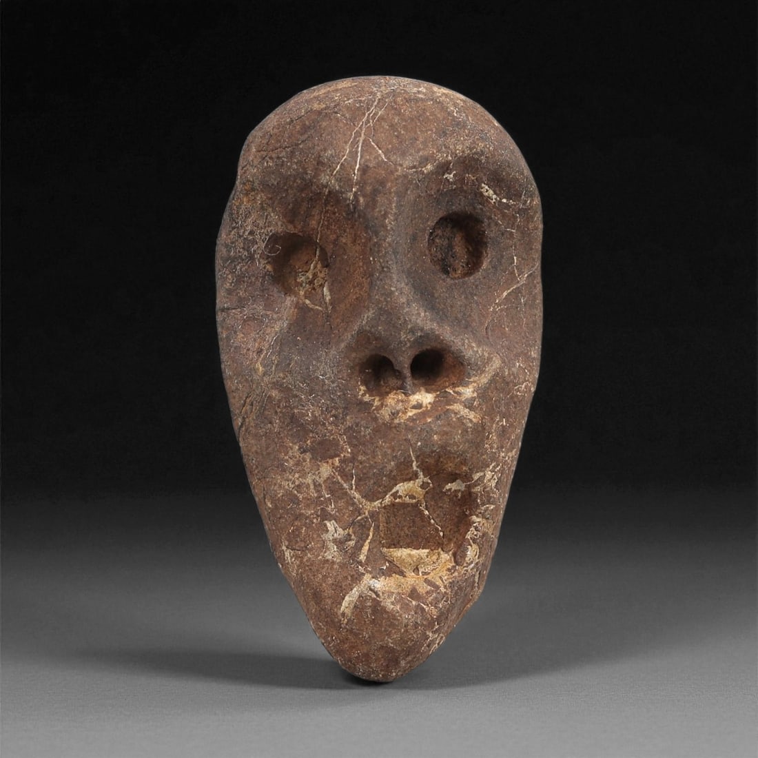 A Mediterranean Stone Maskette, 12th-17th Century, 34AD: Carved from a reddish-brown stone with lighter surface veining and natural fissures, the maskette is shaped as an elongated face tapering to a narrow chin. The eyes are represented by two shallow rece