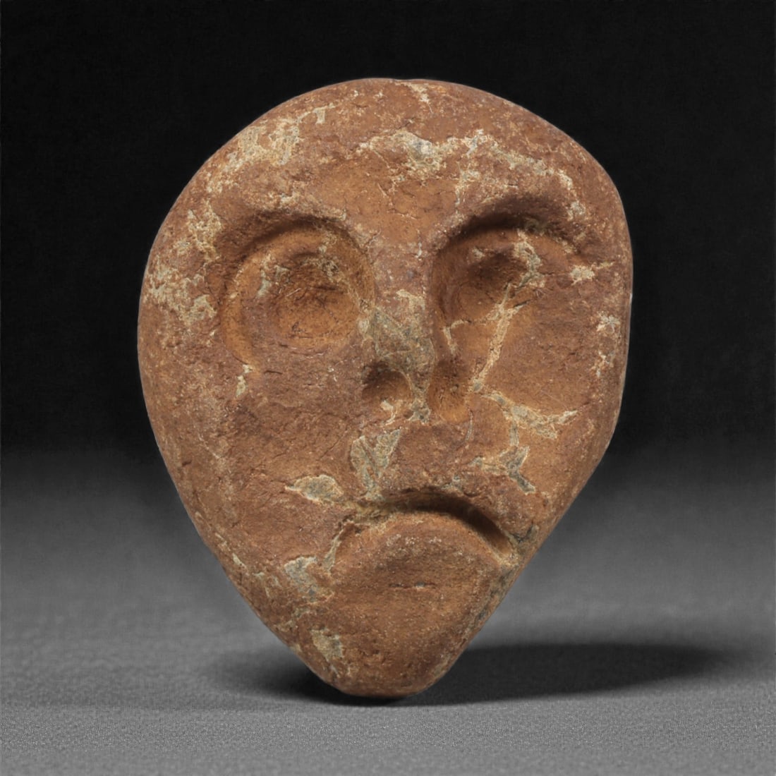 A Mediterranean Stone Maskette, 12th-17th Century, 78AC: Carved from a reddish-brown stone with pale surface veining, the maskette is shaped with an elongated ovoid contour tapering sharply toward the chin. The facial features are rendered in shallow relief