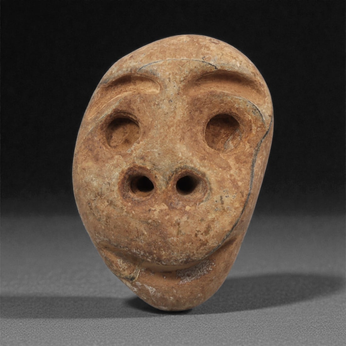 A Mediterranean Stone Maskette, 12th-17th Century, 38AC: Carved from a light brown stone with scattered natural veining, the maskette is shaped with a rounded upper section tapering to a narrow lower edge. The facial features are executed in shallow relief,
