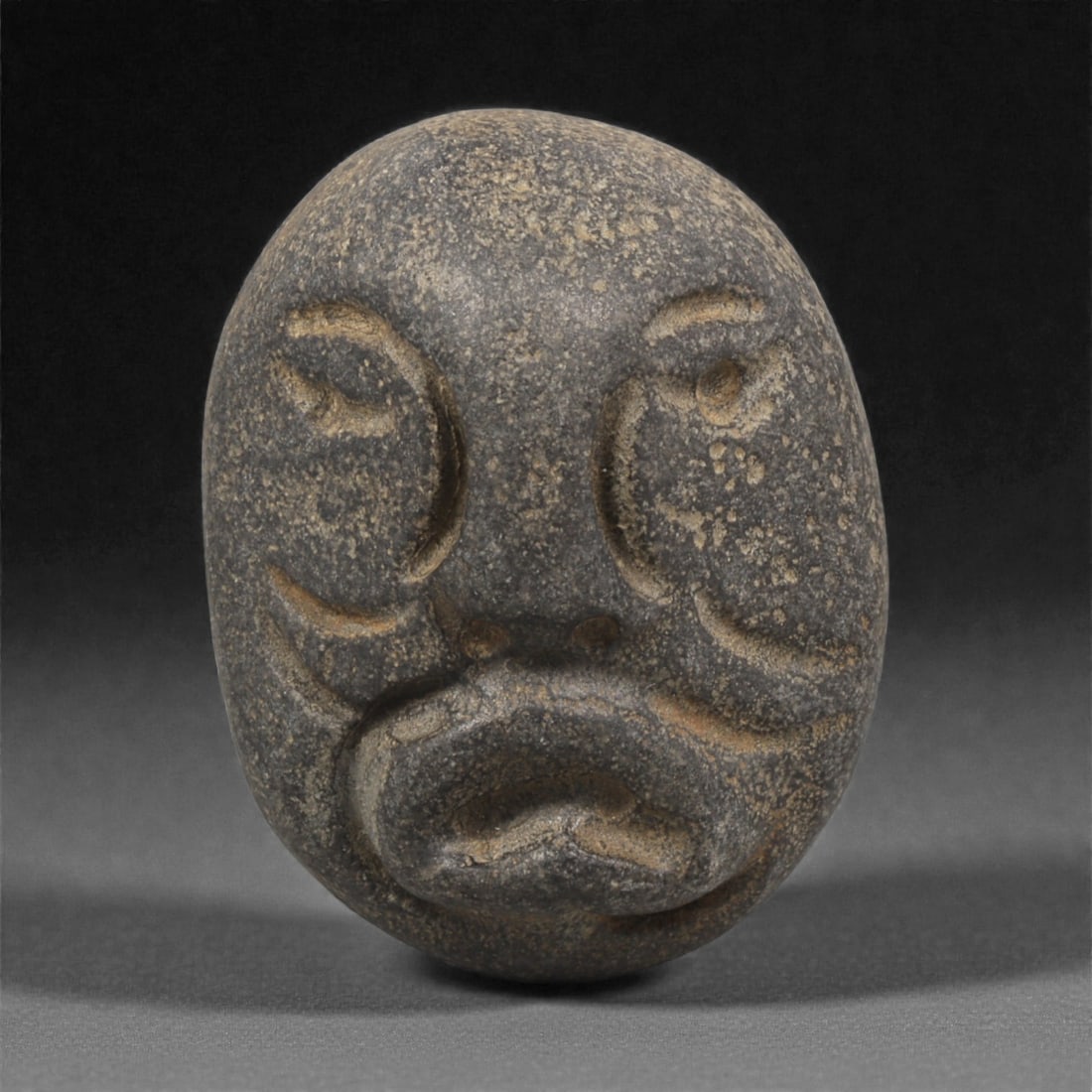 A Mediterranean Stone Maskette, 12th-17th Century, 93AC: Carved from a dark grey stone with lighter granular mottling, the maskette is shaped with an ovoid outline and smoothly rounded contours. The facial features are executed in shallow relief, comprising