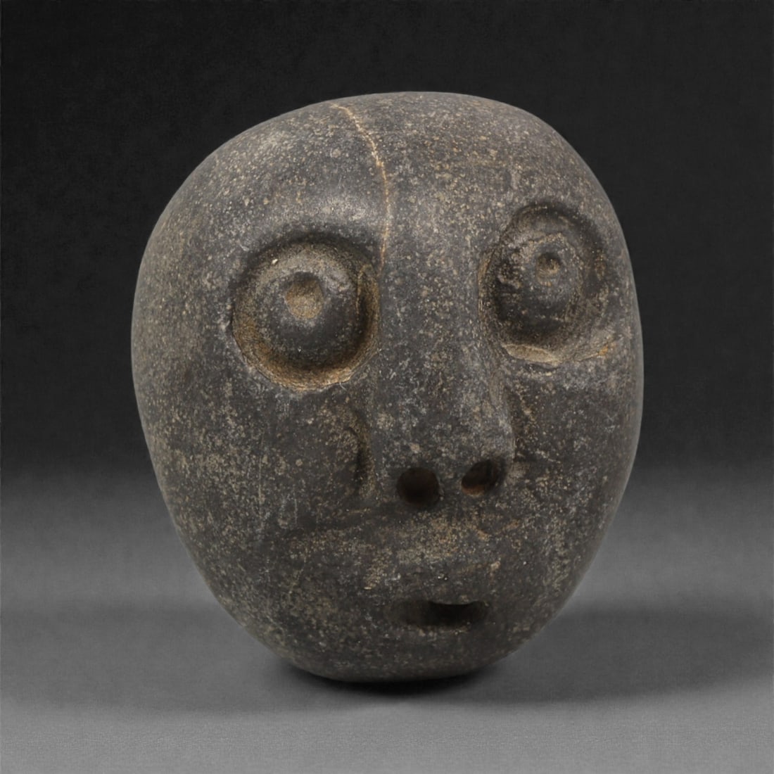 A Mediterranean Stone Maskette, 12th-17th Century, 95AC: Carved from a dark grey stone with fine granular surface texture, the maskette has a rounded cranial outline tapering slightly toward the lower edge. The facial features are rendered in shallow relief