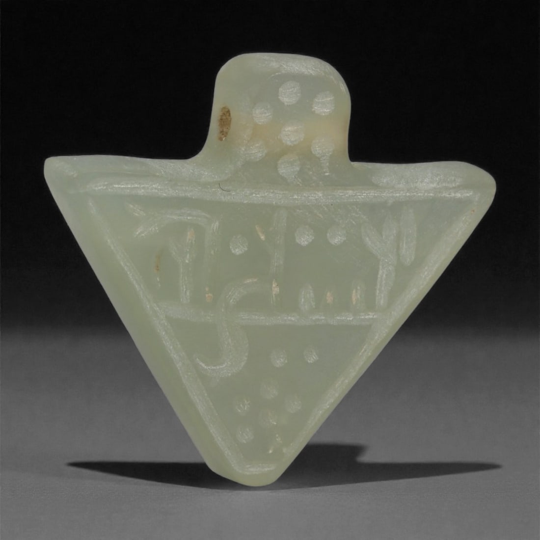 An Islamic Serpentine Jade Triangular Amulet, 10th-13th Century CE, 33LP: Carved from pale green serpentine jade, the amulet is shaped as a small triangular plaque with a rounded upper extension pierced for suspension. The surface bears incised linear motifs and clustered d